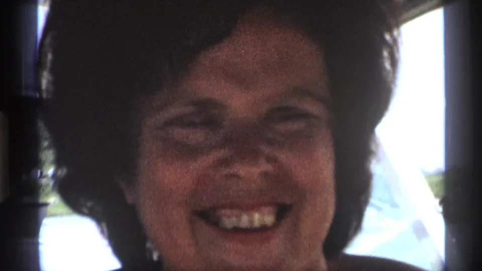The woman smiles broadly in a close home-movie view, with bright window light and soft interior shadows around her.