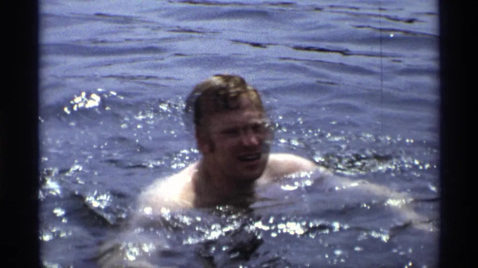 An adult man appears to smile while swimming through blue water, framed in grainy 1973 New York 8mm footage.