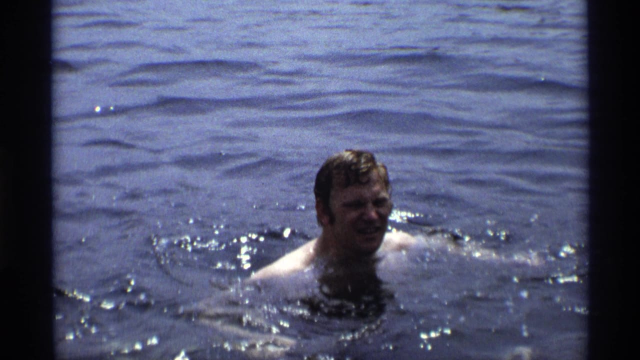 A man swims in lake water during a 1973 New York home-movie family outing, with ripples and bright reflections around him.