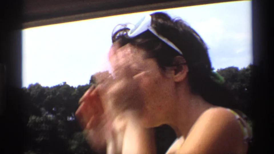 A woman in a swimsuit turns or laughs near the lake, her sunglasses resting on her head, with trees and sky behind her.