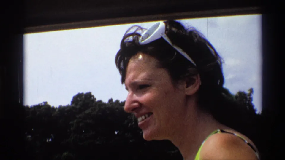 A smiling woman in profile appears near a lake with sunglasses on her head, backed by trees and sky in 1973 New York footage.
