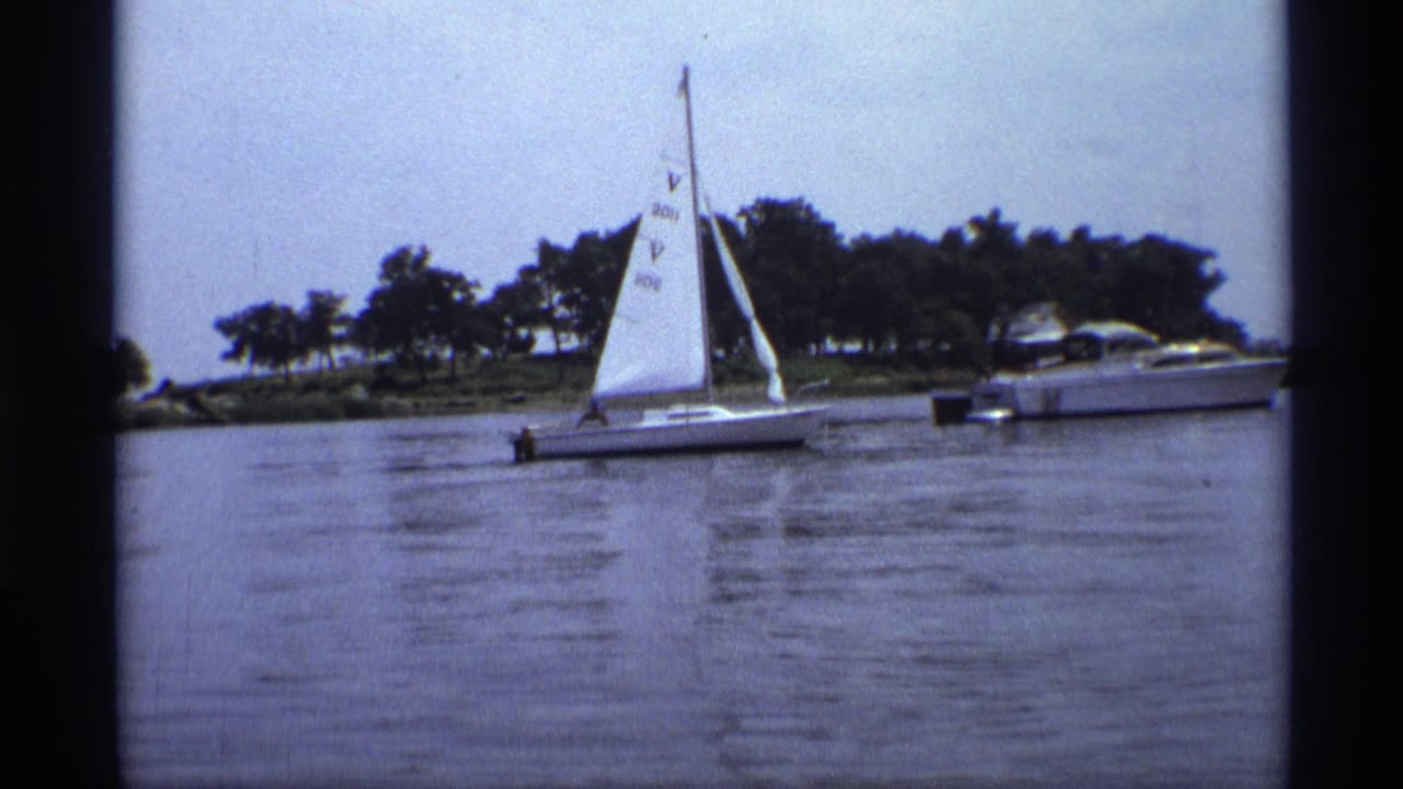 Small sailboat crossing calm water with a motorboat and tree-lined shore behind it.