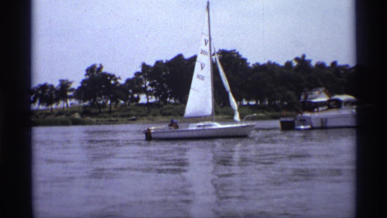1973 home-movie frame of a sailboat on a New York lake, with soft film grain and a motorboat near the wooded bank.
