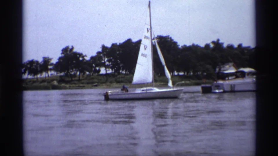1973 home-movie frame of a sailboat on a New York lake, with soft film grain and a motorboat near the wooded bank.