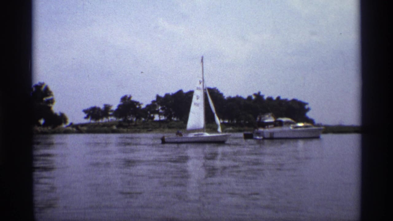 Wide 1973 New York lake view with a small sailboat, nearby motorboat, calm water, and trees under a bright sky.