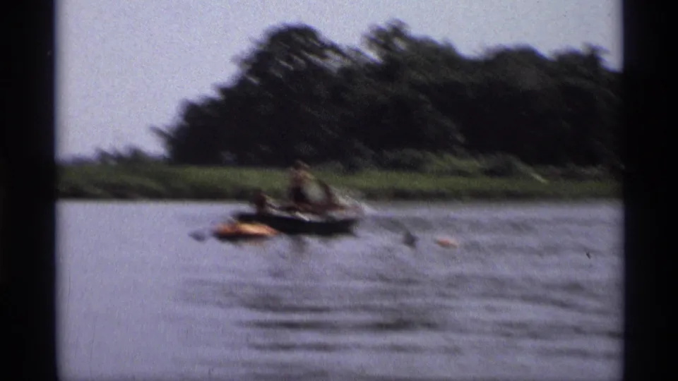 1973 New York 8mm home-movie frame showing a distant small boat and orange raft on calm lake water near a wooded shoreline.