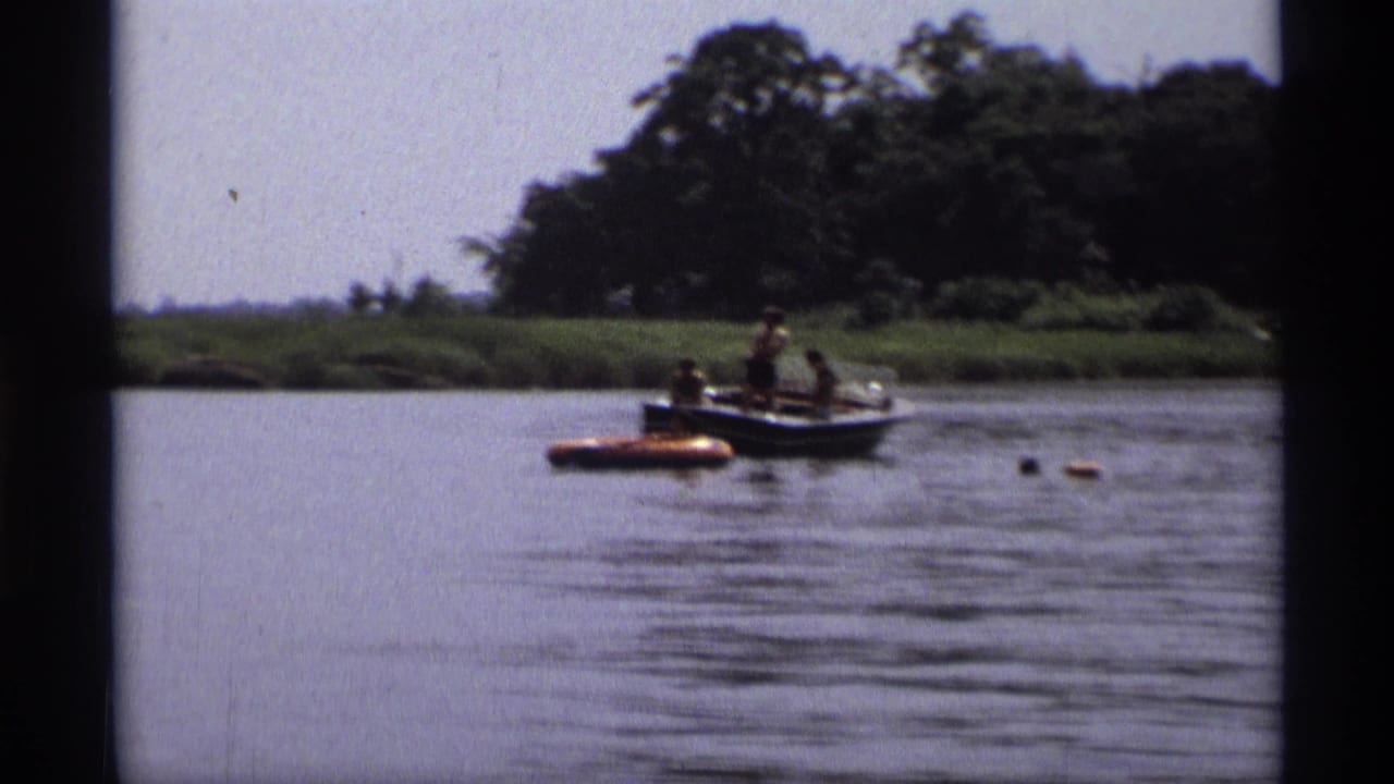 Distant 1973 New York lake scene with several people in a small boat beside an orange inflatable raft and wooded shoreline.