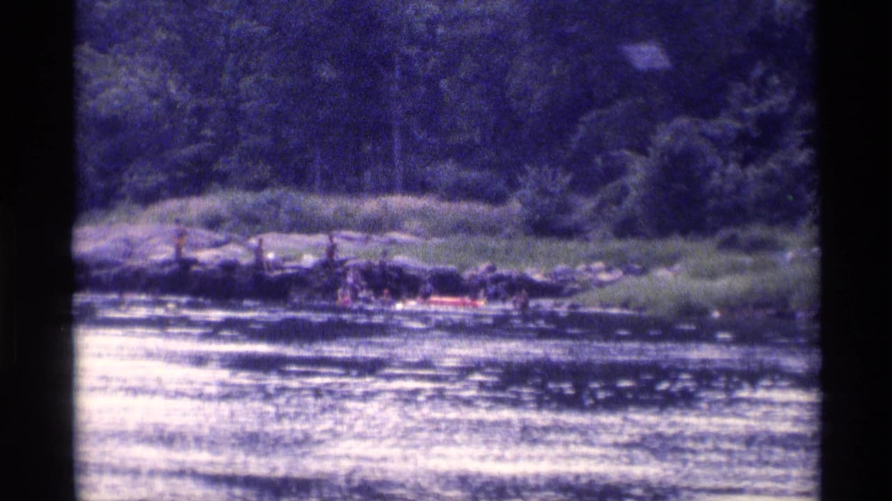 Distant 8mm frame of a wooded New York lake in 1973, showing people near rocks and a red inflatable raft.