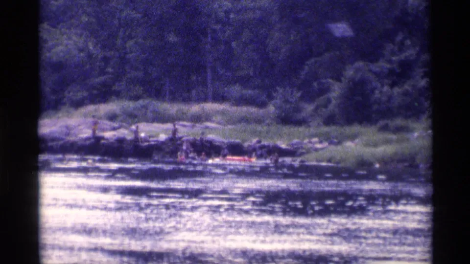 Distant 8mm frame of a wooded New York lake in 1973, showing people near rocks and a red inflatable raft.
