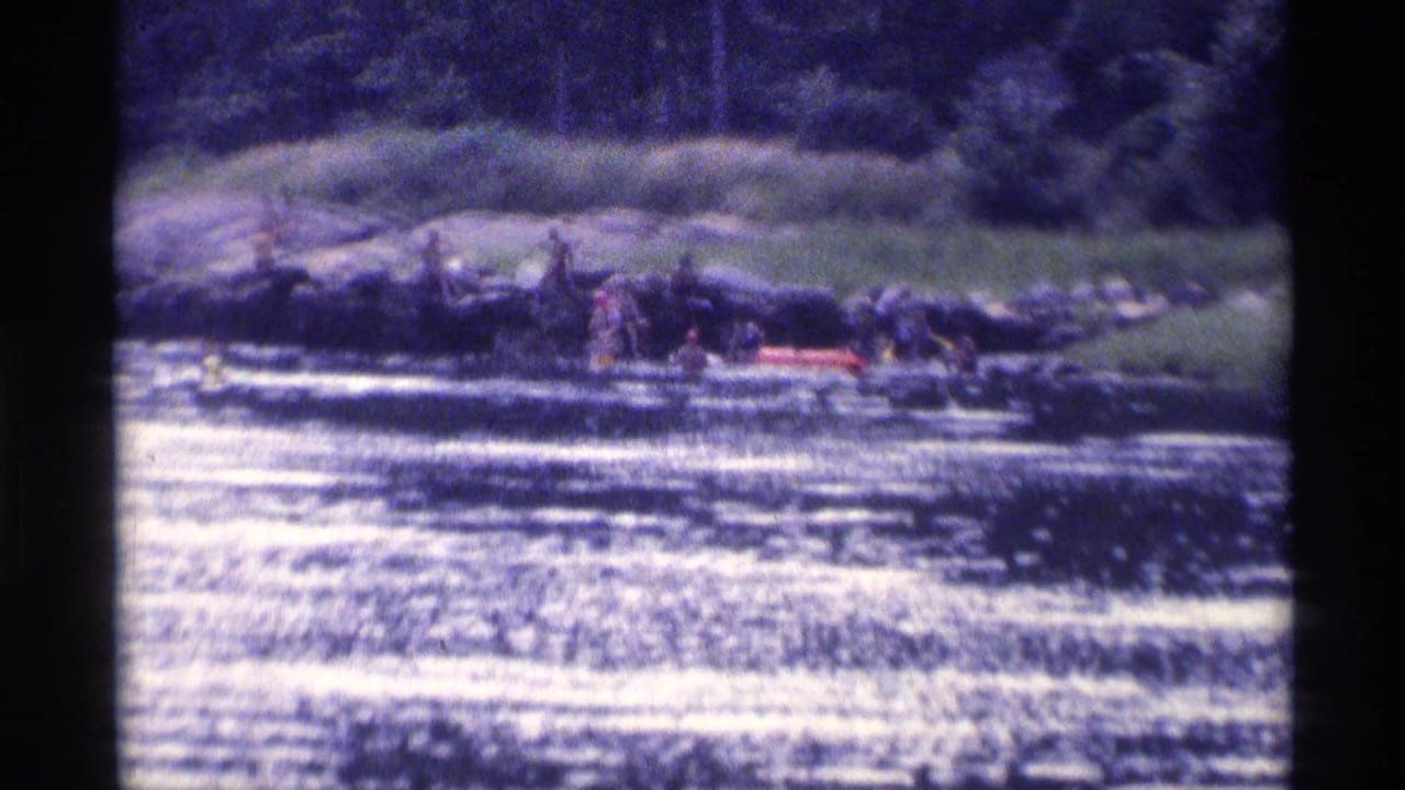 1973 home-movie frame showing young men at a wooded New York lakeshore with a red raft beside the rocks.