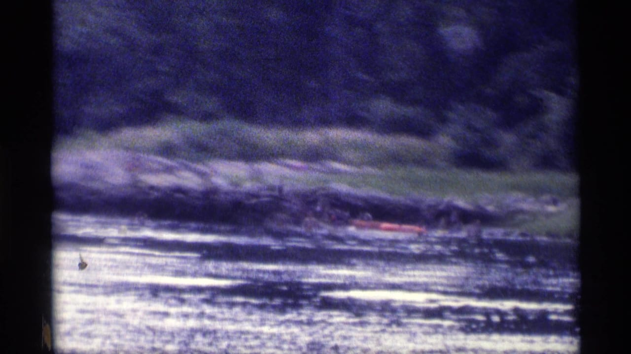 Soft, grainy 8mm view of a New York lake shoreline in 1973, with people and a red raft partly visible.