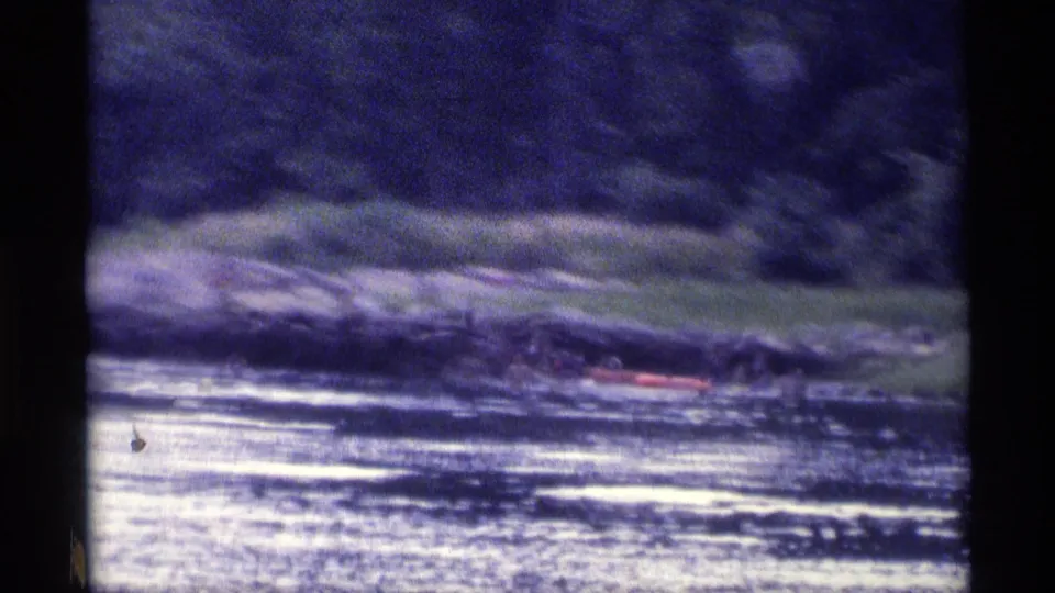 Soft, grainy 8mm view of a New York lake shoreline in 1973, with people and a red raft partly visible.