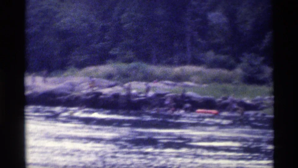 1973 New York 8mm frame showing rippled lake water, a rocky tree-lined shore, and a small red boat near the bank.