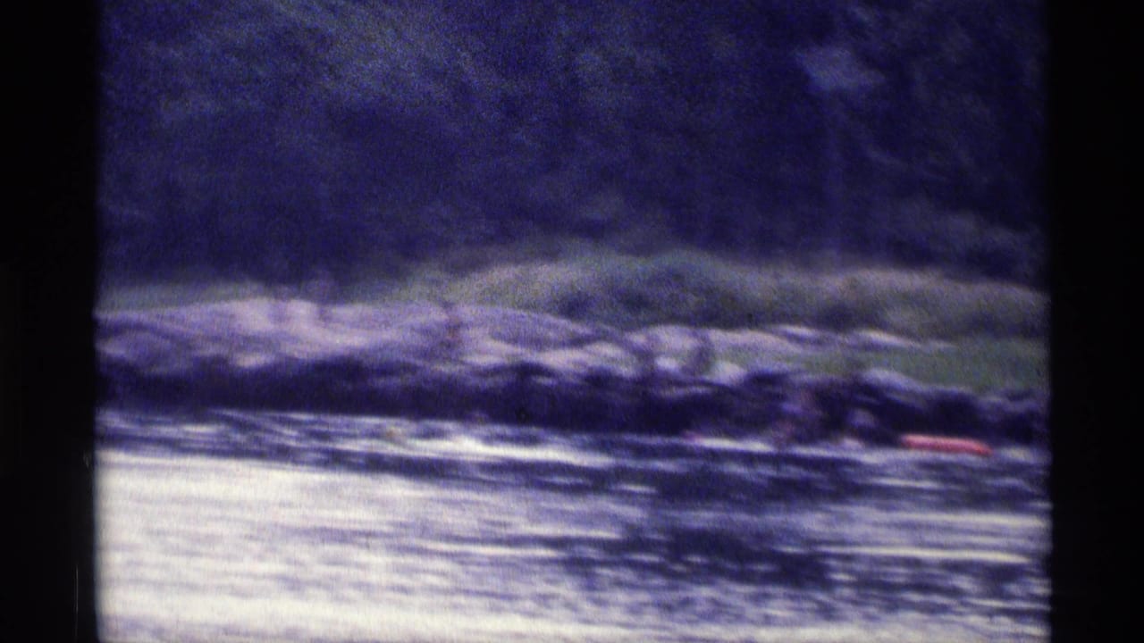 Distant 1973 New York lake view with textured water, dark trees, rocky shore, and a red boat along the shoreline.