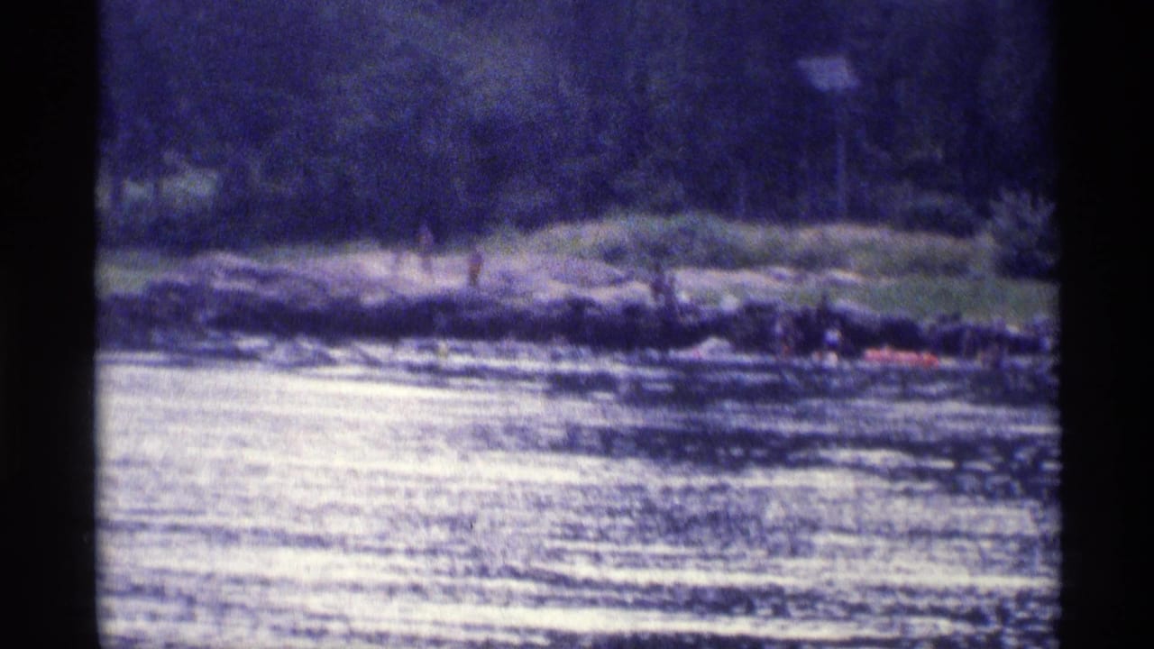 1973 New York lakeside frame showing people gathered along a rocky shore, with rippling water, trees, and a red boat nearby.
