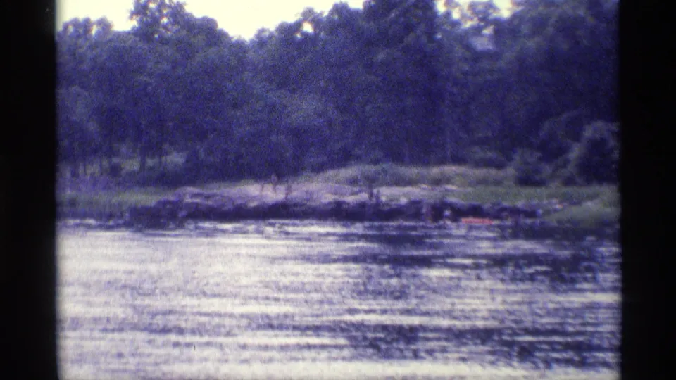 1973 New York 8mm frame of calm water facing a tree-lined bank, with soft focus and blue-tinted film color.
