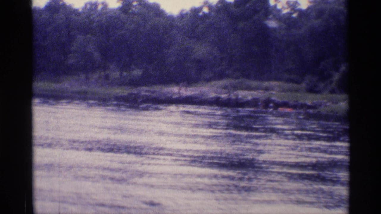 1973 New York 8mm view of a lake or river edge, with trees, rippled water, and possible distant people near the shoreline.