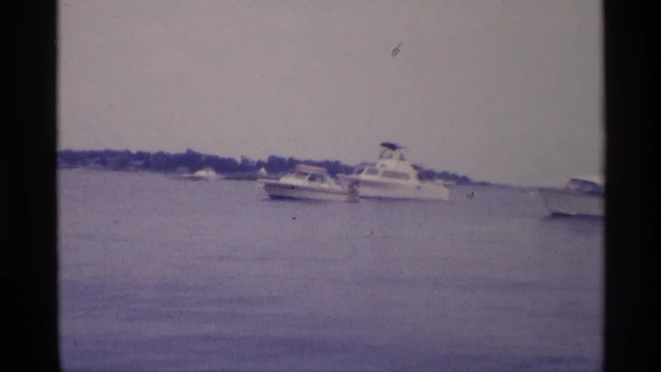 1973 New York 8mm frame showing white motorboats on calm basin water beneath an overcast sky, with a distant tree-lined shoreline.