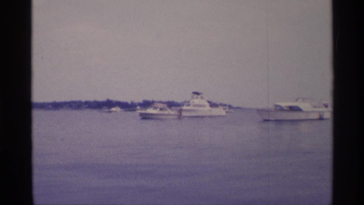 Soft 8mm view of a cabin cruiser and smaller pleasure boats floating on calm basin water in New York, 1973.
