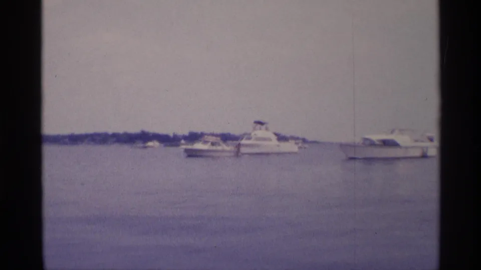 Soft 8mm view of a cabin cruiser and smaller pleasure boats floating on calm basin water in New York, 1973.
