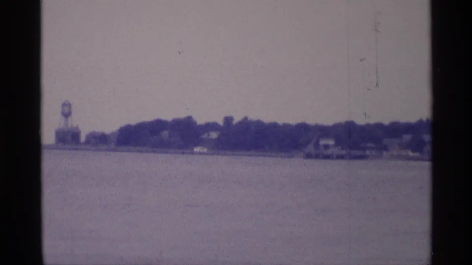 1973 New York 8mm frame showing a hazy shoreline across open water, with a distant water tower and low buildings under cloudy sky.