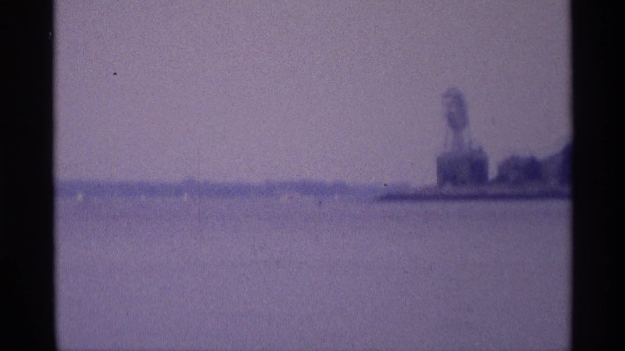 Soft 8mm view across a broad water surface toward a distant shoreline water tower in New York, tinted purple-blue by the film scan.