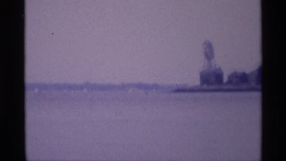 Soft 8mm view across a broad water surface toward a distant shoreline water tower in New York, tinted purple-blue by the film scan.