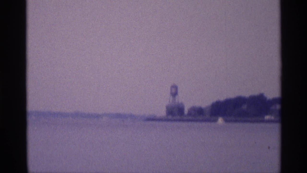 1973 New York home-movie frame with open water in the foreground and a distant water tower beside a low, tree-lined shore.