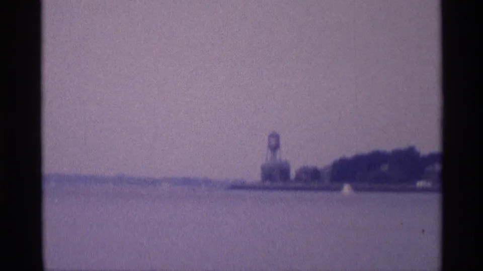 1973 New York home-movie frame with open water in the foreground and a distant water tower beside a low, tree-lined shore.