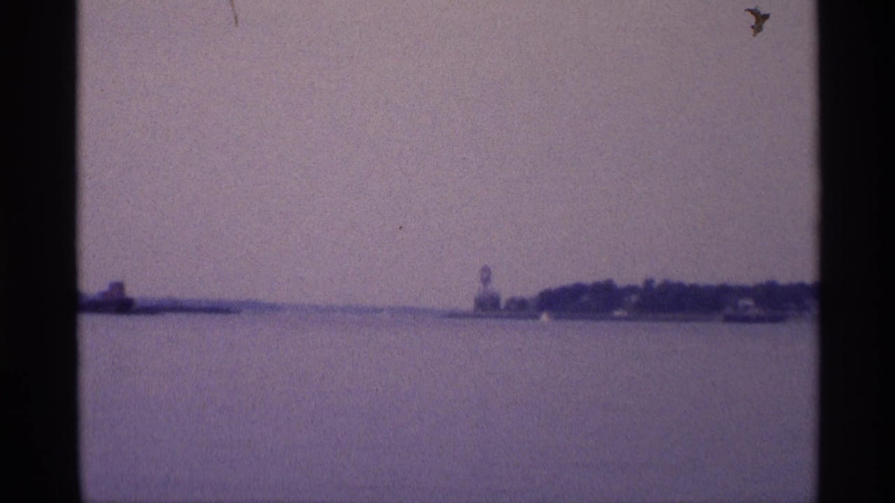 Wide 1973 New York waterfront frame showing open water, a low distant shoreline, and a small water tower under a cloudy sky.