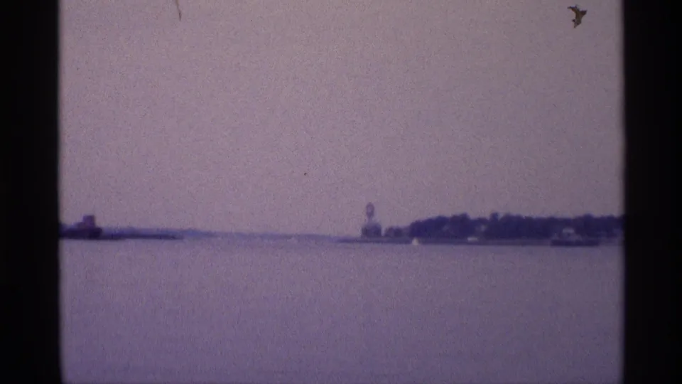 Wide 1973 New York waterfront frame showing open water, a low distant shoreline, and a small water tower under a cloudy sky.