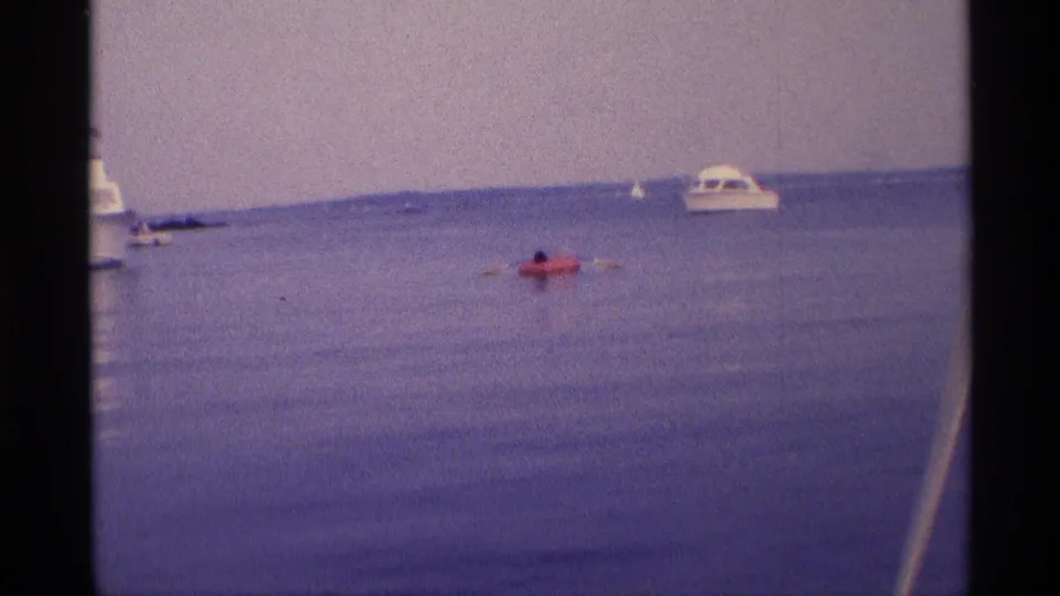 1973 New York 8mm frame with a distant person paddling a red inflatable raft near a white motorboat and small boats.