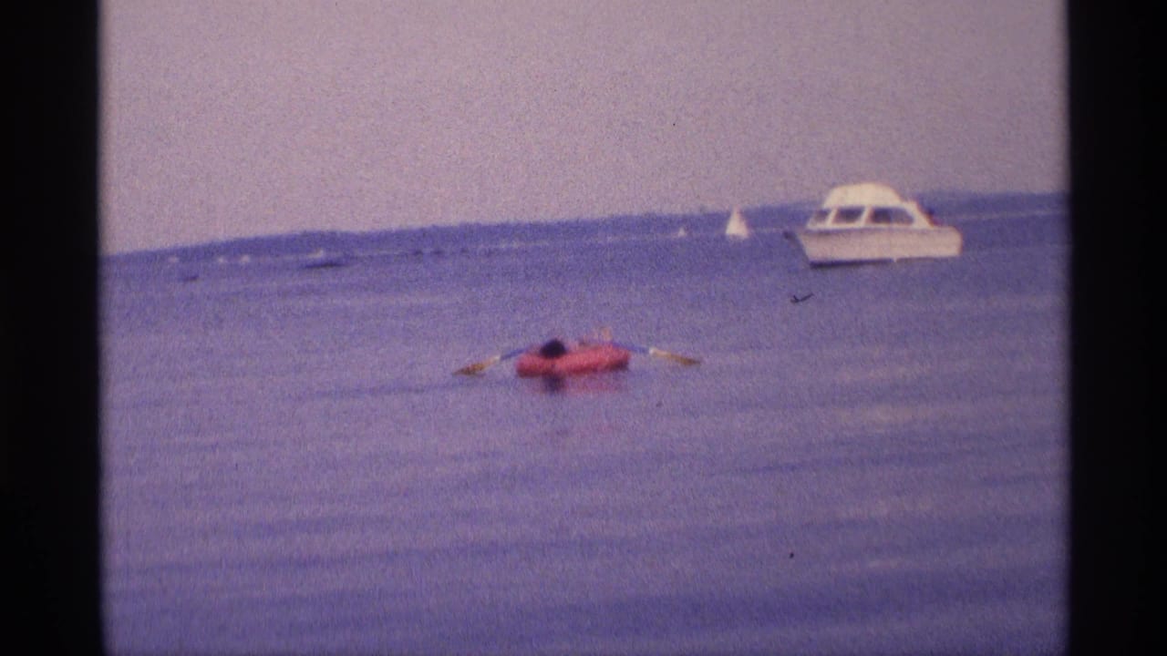 1973 New York 8mm frame showing a person paddling a red inflatable raft across calm water near a white cabin motorboat.