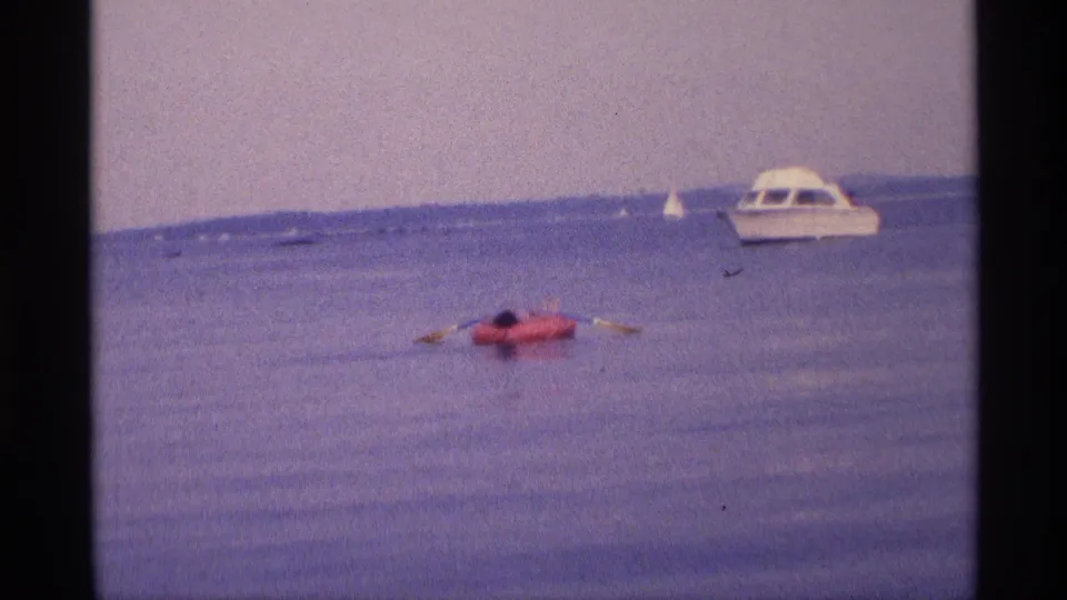 1973 New York 8mm frame showing a person paddling a red inflatable raft across calm water near a white cabin motorboat.