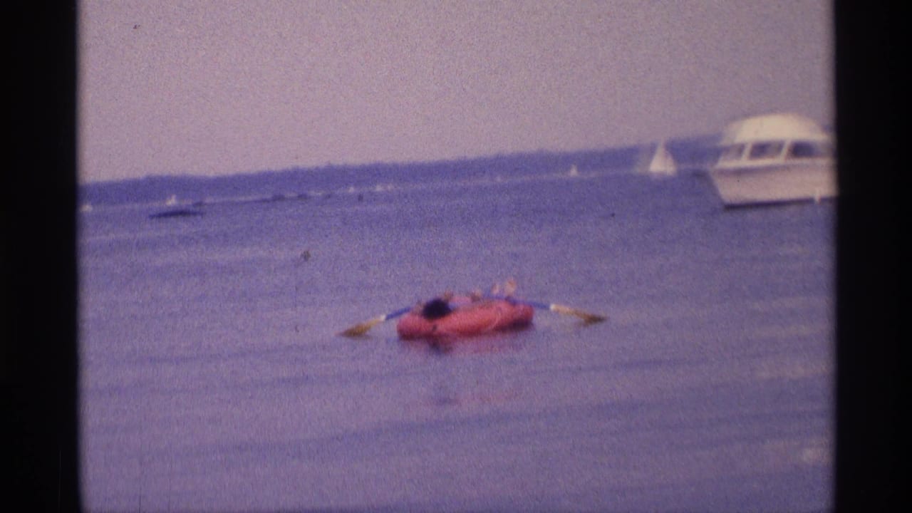 1973 New York 8mm frame with a red inflatable raft, paddler, oars, distant sailboats, and a white motorboat near the edge.