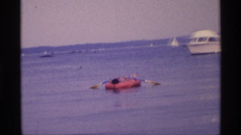 1973 New York 8mm frame with a red inflatable raft, paddler, oars, distant sailboats, and a white motorboat near the edge.