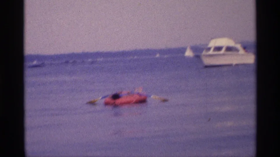 1973 New York 8mm frame showing a woman drifting on a red raft near a white motorboat on open water.