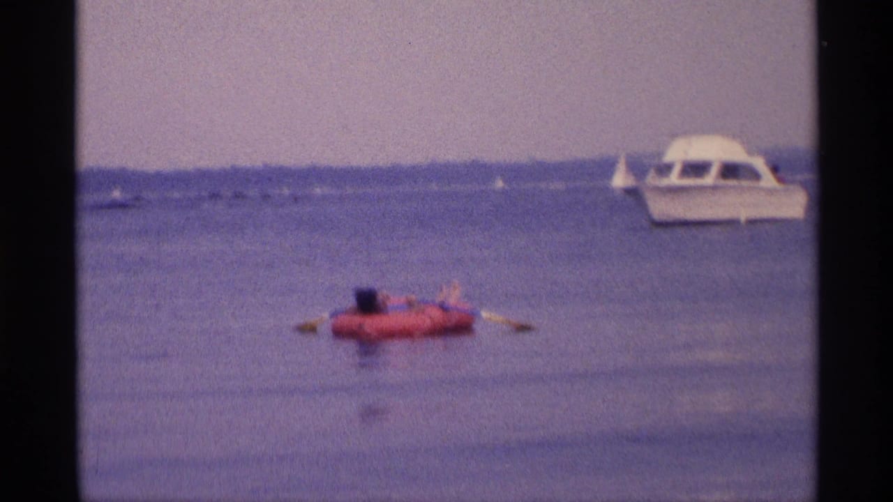 1973 New York home-movie frame of a woman on a red raft, with small boats and a white motorboat in the distance.