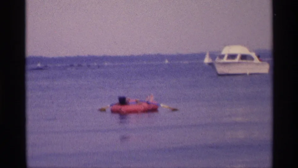 1973 New York home-movie frame of a woman on a red raft, with small boats and a white motorboat in the distance.