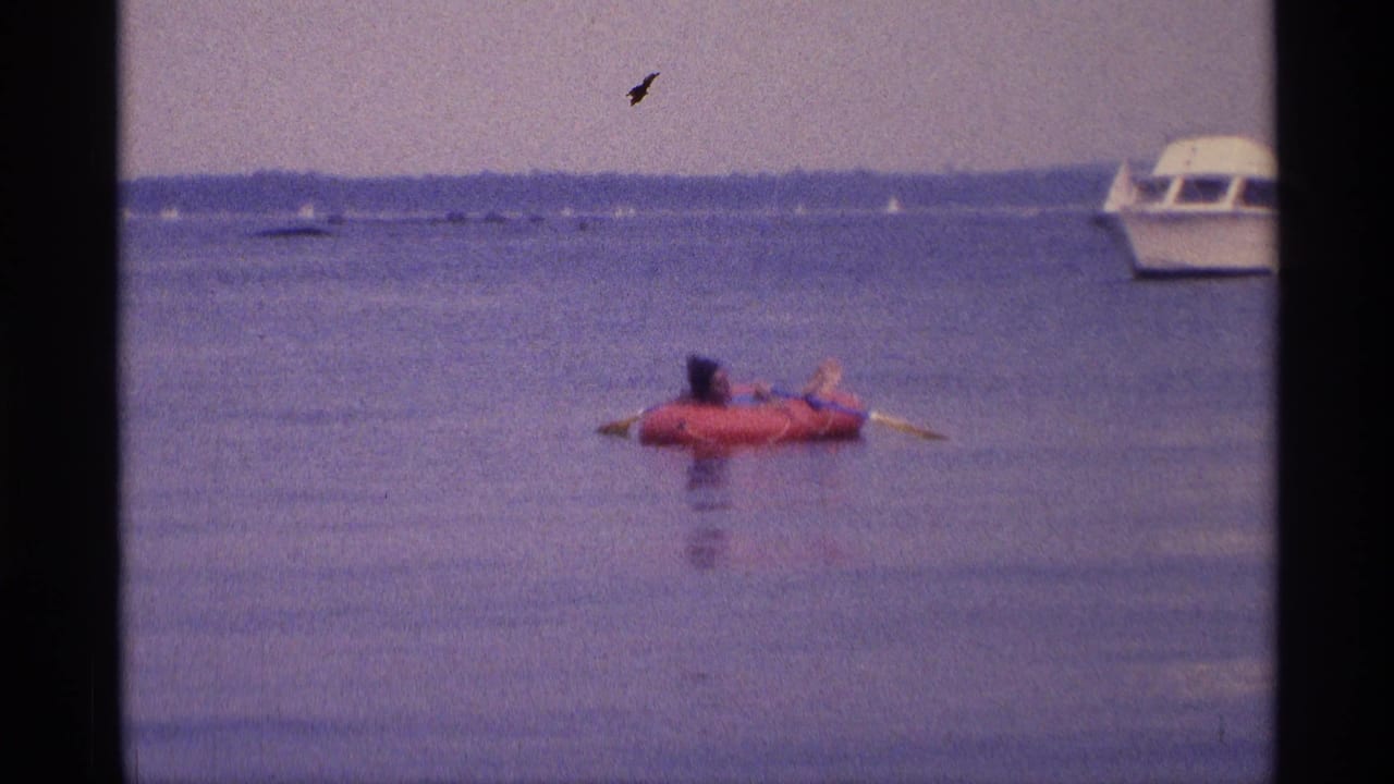 Soft 8mm frame from 1973 New York showing a woman on a red raft, distant boats, and a small film mark in the sky.