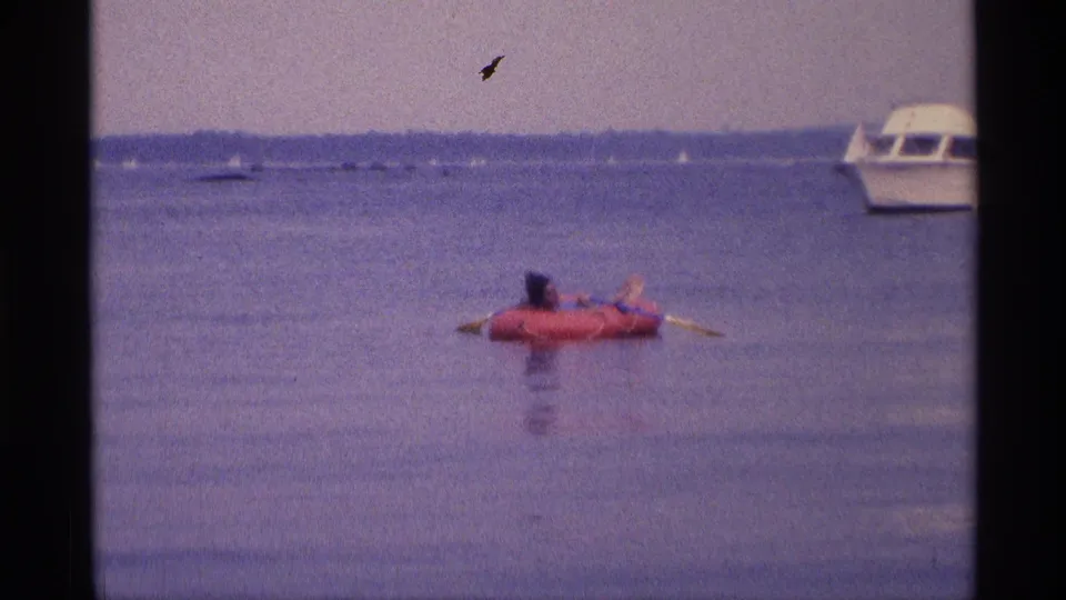 Soft 8mm frame from 1973 New York showing a woman on a red raft, distant boats, and a small film mark in the sky.