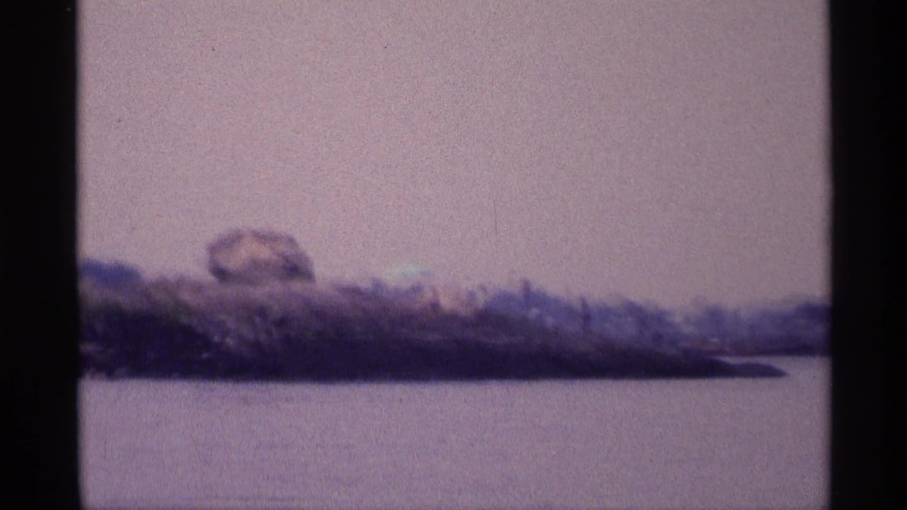 Calm lake with rocky shoreline, boulders, low greenery, and pale sky.