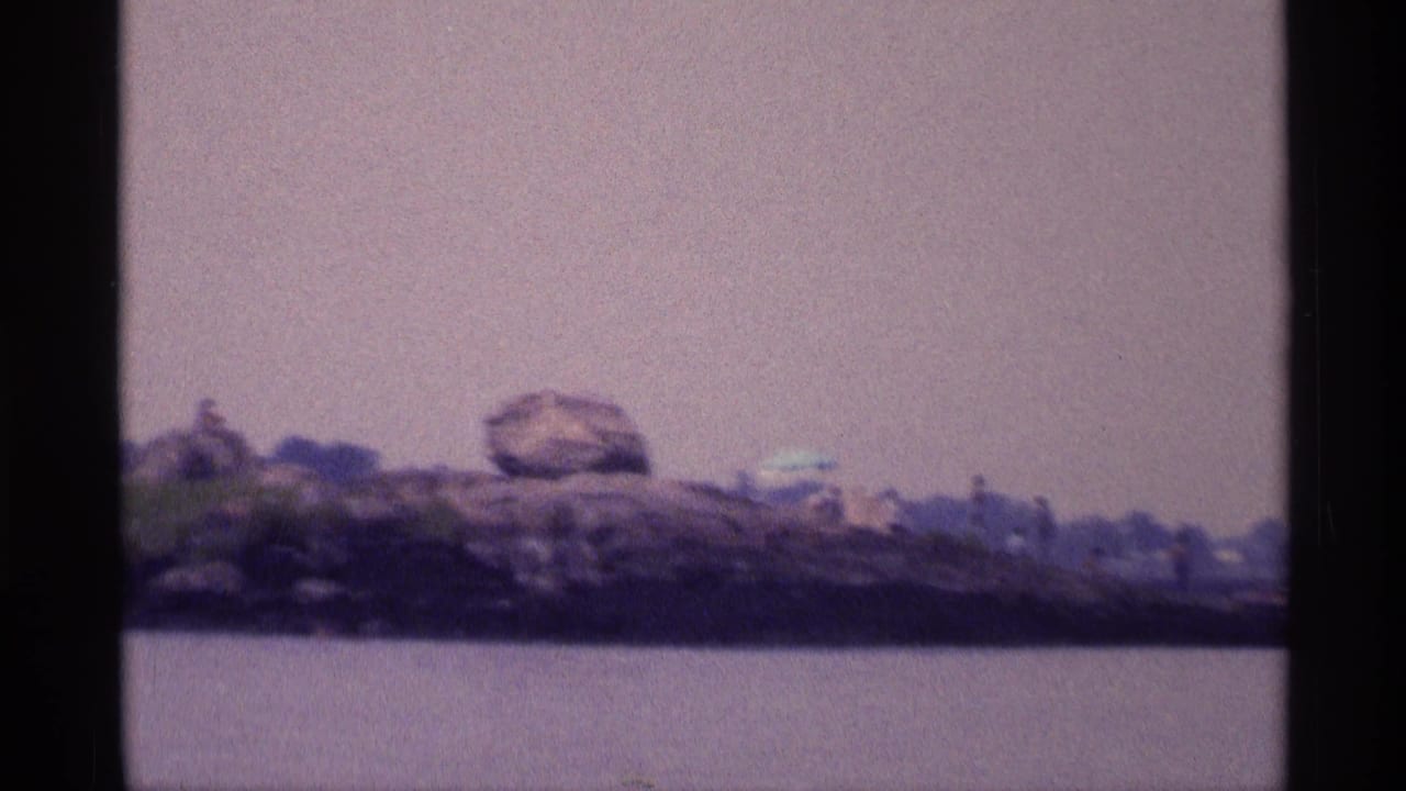 Rocky shore across the lake with boulders, greenery, and tiny distant figures.