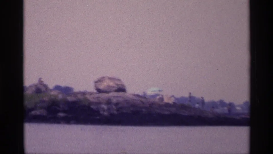 1973 New York 8mm frame with a rocky lakeside bank, large boulders, sparse greenery, and small distant people.