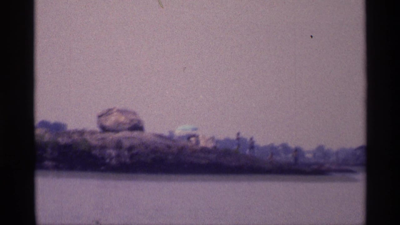 1973 New York 8mm frame looking over water toward a rocky shore with a prominent boulder and faded green brush.
