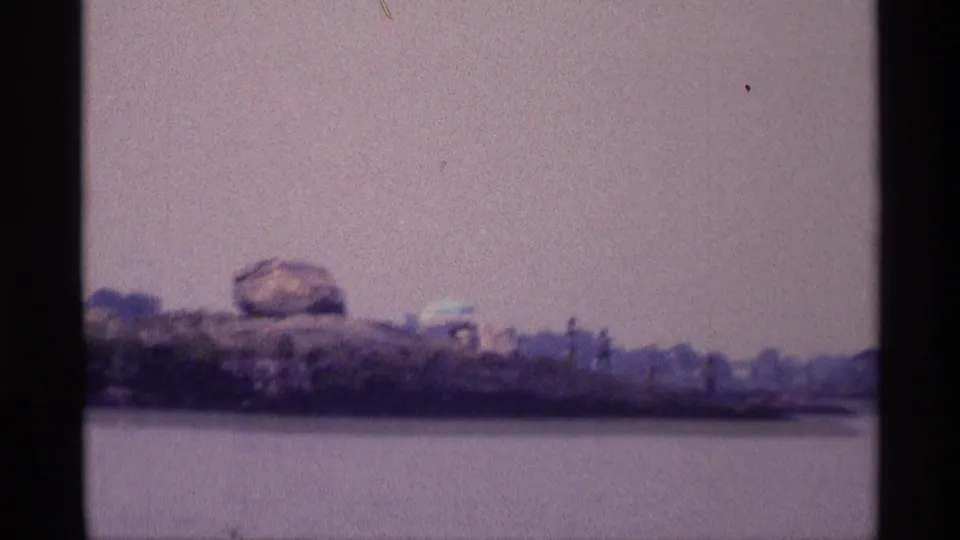 1973 New York 8mm frame looking over water toward a rocky shore with a prominent boulder and faded green brush.