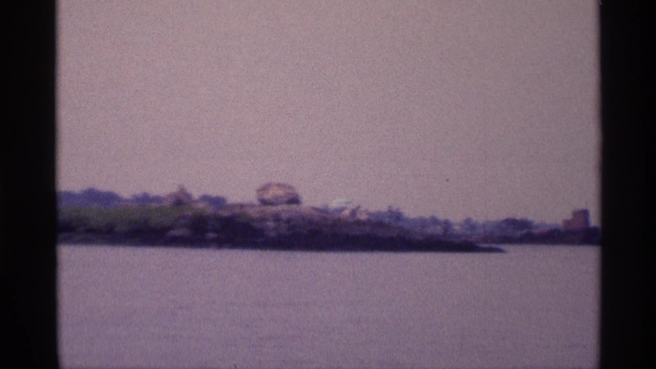 1973 New York 8mm frame showing a wider lake view with rocky shoreline, boulders, sparse greenery, and tiny people in the distance.