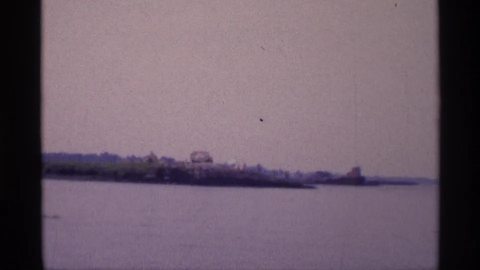 1973 New York 8mm frame showing calm water and a low, distant shoreline under a muted hazy sky.