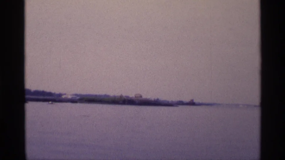 1973 New York waterfront view with open water, a low tree-lined shore, and soft purple-blue 8mm color.