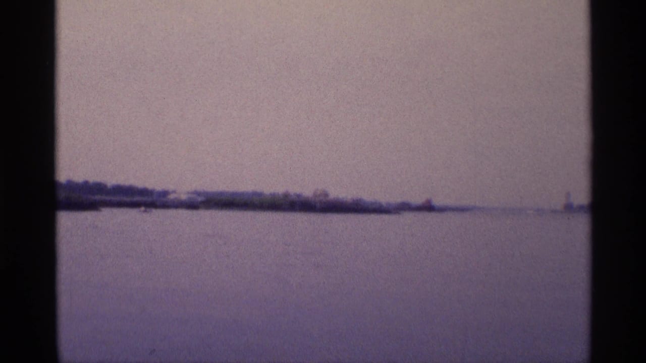 Faded 1973 New York 8mm frame with calm water, low shoreline, and a small distant vertical structure near the horizon.